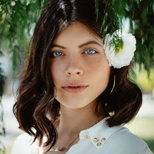 Close-up portrait of a woman with blue eyes and dark hair wearing a white flower outdoors.